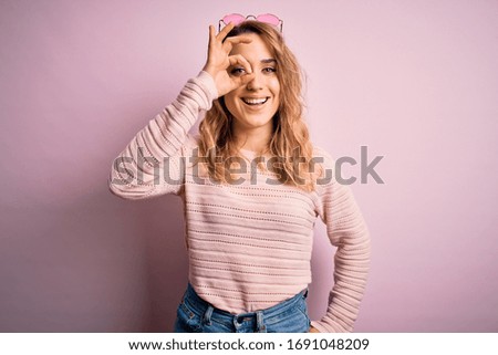 Young beautiful blonde woman wearing casual sweater and sunglasses over pink background doing ok gesture with hand smiling, eye looking through fingers with happy face.