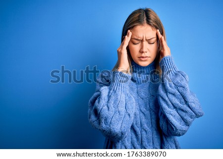 Young beautiful blonde woman wearing winter wool sweater over blue isolated background with hand on head for pain in head because stress. Suffering migraine.