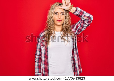 Young beautiful blonde woman wearing casual shirt standing over isolated red background making fun of people with fingers on forehead doing loser gesture mocking and insulting.