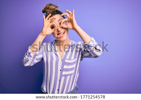 Young beautiful blonde woman wearing casual striped shirt standing over purple background doing ok gesture like binoculars sticking tongue out, eyes looking through fingers. Crazy expression.