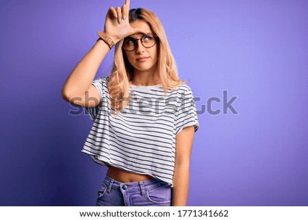 Young beautiful blonde woman wearing striped t-shirt and glasses over purple background making fun of people with fingers on forehead doing loser gesture mocking and insulting.