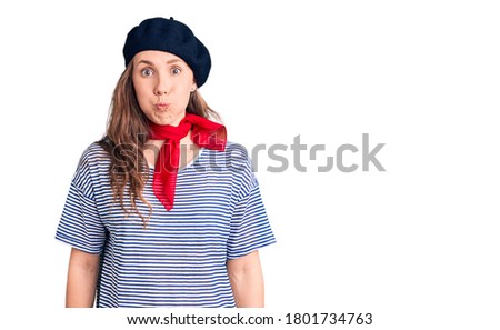 Young beautiful blonde woman wearing french beret and striped t-shirt puffing cheeks with funny face. mouth inflated with air, crazy expression. 