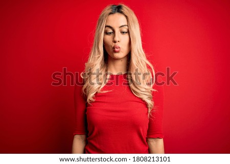 Young beautiful blonde woman wearing casual t-shirt standing over isolated red background making fish face with lips, crazy and comical gesture. Funny expression.