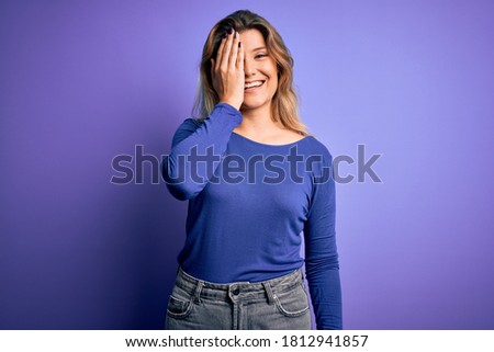 Young beautiful blonde woman wearing casual t-shirt over isolated purple background covering one eye with hand, confident smile on face and surprise emotion.