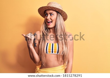 Young beautiful blonde woman wearing bikini and summer hat over isolated yellow background pointing thumb up to the side smiling happy with open mouth