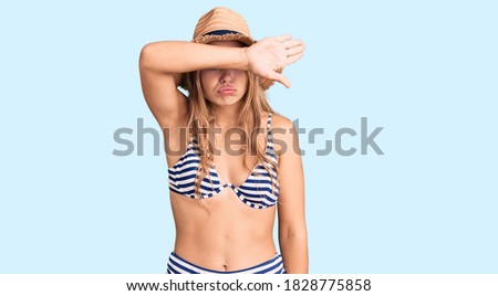 Young beautiful blonde woman wearing bikini and hat covering eyes with arm, looking serious and sad. sightless, hiding and rejection concept 
