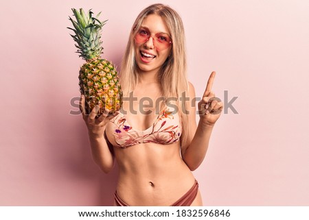 Young beautiful blonde woman wearing bikini and sunglasses holding pineapple smiling with an idea or question pointing finger with happy face, number one 