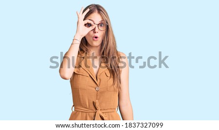 Young beautiful blonde woman wearing casual clothes and glasses doing ok gesture shocked with surprised face, eye looking through fingers. unbelieving expression. 