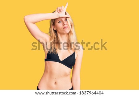 Young beautiful blonde woman wearing bikini making fun of people with fingers on forehead doing loser gesture mocking and insulting. 