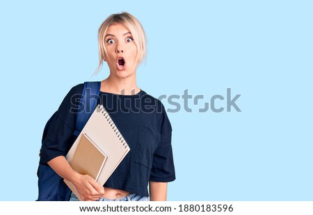 Young beautiful blonde woman wearing student backpack holding notebook scared and amazed with open mouth for surprise, disbelief face 