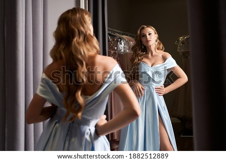 Young beautiful blonde woman wearing an off-the-shoulder full-length sky blue satin slit prom ball gown. Model looking in mirror.