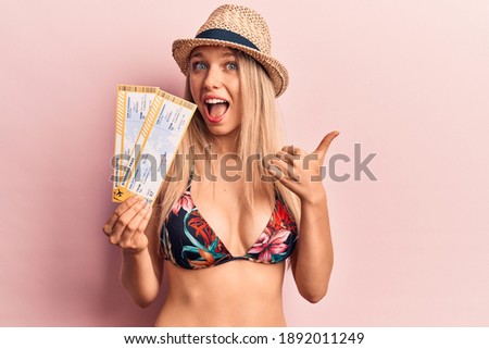 Young beautiful blonde woman wearing bikini and hat holding boarding pass pointing thumb up to the side smiling happy with open mouth 