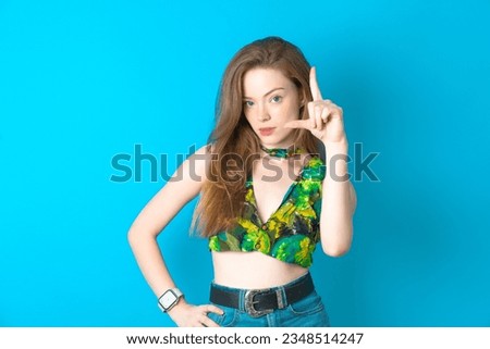 young beautiful blonde woman wearing green top standing over blue studio background making fun of people with fingers on forehead doing loser gesture mocking and insulting.