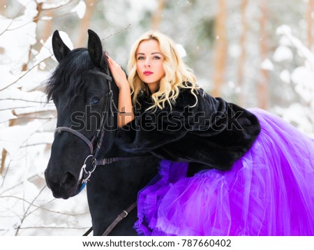 Young beautiful blonde woman wearing a long pink dress riding a black horse in the snowy winter forest
