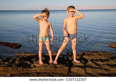 Young beautiful boy and girl stay in front of water looking forward with hands near eyes. Barefoot on rocks and reefs, children stand looking for amazing future.Two friends are happy to play in summer