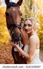  A young beautiful bride in a white dress stands near a horse in the autumn forest