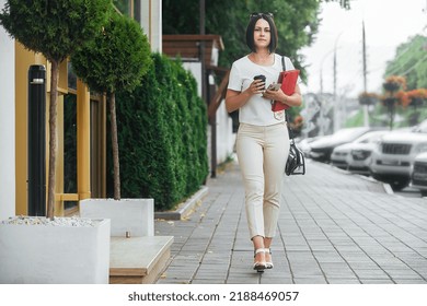 a young beautiful brunette business woman in a white blouse and light trousers walks along the city street, in her hands a glass of coffee and a tablet