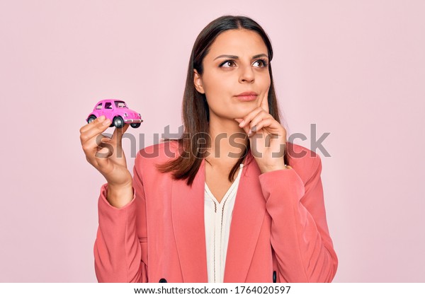 Young beautiful brunette\
business woman holding small car toy over pink background serious\
face thinking about question with hand on chin, thoughtful about\
confusing idea