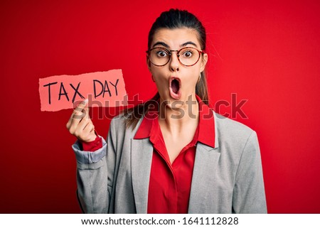 Young beautiful brunette businesswoman holding papaer with tax day message scared in shock with a surprise face, afraid and excited with fear expression