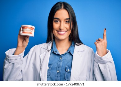 Young beautiful brunette dentist woman wearing coat holding plastic teeth prosthesis surprised with an idea or question pointing finger with happy face, number one