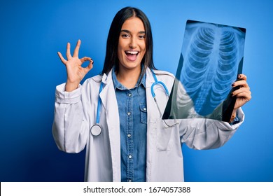 Young beautiful brunette doctor woman wearing stethoscope holding chest xray doing ok sign with fingers, excellent symbol