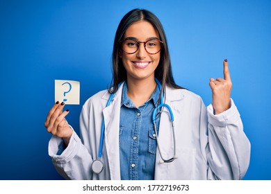 Young Beautiful Brunette Doctor Woman Holding Paper With Question Mark Symbol Message Surprised With An Idea Or Question Pointing Finger With Happy Face, Number One