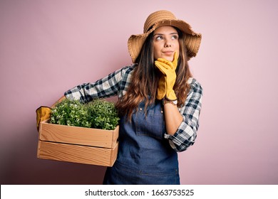 Young Beautiful Brunette Farmer Woman Wearing Apron And Hat Holding Box With Plants Serious Face Thinking About Question, Very Confused Idea