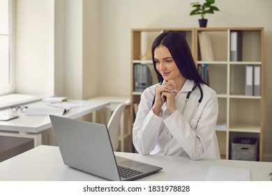 Young Beautiful Brunette Female Doctor Sitting In The Office In Front Of A Laptop And Reviewing The Results Of The Examination For The Disease. Smiling Woman Is Happy That Her Patient Is Healthy.