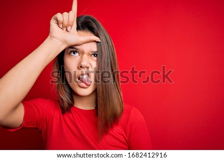 Young beautiful brunette girl wearing casual t-shirt over isolated red background making fun of people with fingers on forehead doing loser gesture mocking and insulting.