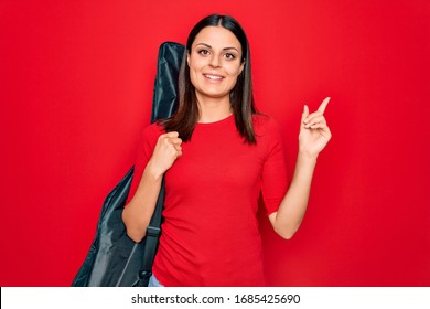 Young Beautiful Brunette Guitarist Woman Holding Guitar Case Over Isolated Red Background Smiling Happy Pointing With Hand And Finger To The Side