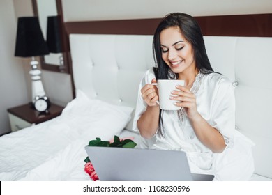 The young beautiful brunette happy woman lies in her bed in the morning. The girl looks at the laptop, she laughs and drinks tea from the cute cup with big bouquet of roses.