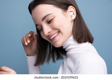 A young and beautiful brunette listens to music on wireless headphones in the studio on a blue background. Close-up portrait.