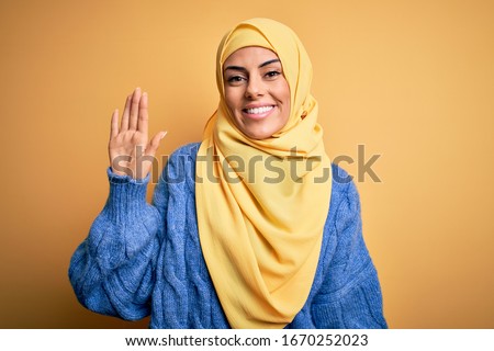 Young beautiful brunette muslim woman wearing arab hijab over isolated yellow background Waiving saying hello happy and smiling, friendly welcome gesture