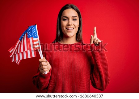 Young beautiful brunette patriotic woman holding united states flag over red background surprised with an idea or question pointing finger with happy face, number one