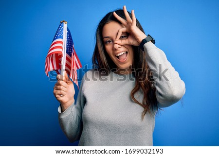 Young beautiful brunette patriotic woman holding american flag celebrating 4th of july with happy face smiling doing ok sign with hand on eye looking through fingers