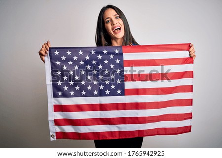 Young beautiful brunette patriotic woman with blue eyes smiling happy and confident. Standing with smile on face holding united states flag celebrating independence day over isolated white background
