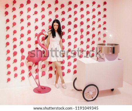 Young beautiful brunette posing against a wall background with pink ice cream and equipment for making sweet cotton wool. Sweet decorations on the background