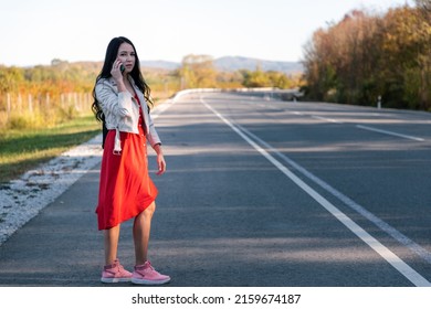 A young beautiful brunette in a red dress hitchhiking and calling on the phone. In the background, the road goes into the distance and the trees. Copy space. The concept of hitchhiking.