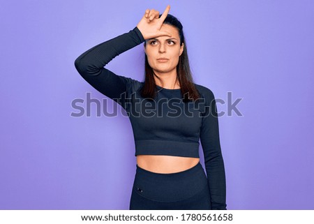 Young beautiful brunette sporty woman wearing casual sportswear over purple background making fun of people with fingers on forehead doing loser gesture mocking and insulting.