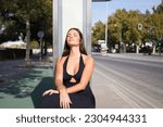 Young beautiful brunette typical Spanish woman dressed in black suit sitting at bus stop waiting. The woman is happy and relaxed under the sun of seville. Public transport concept.