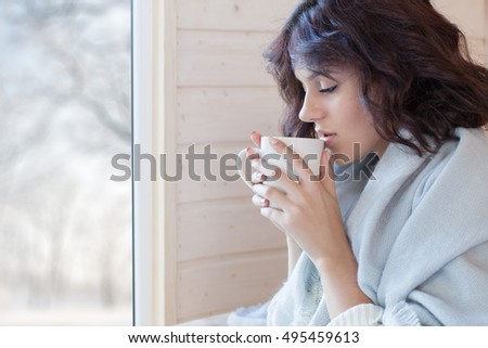 Young beautiful brunette woman covered in blanket with cup of coffee sitting home by the window. Winter snow landscape view. Lazy day off concept
