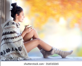 Young Beautiful  Brunette Woman With Cup Of Coffee Wearing Knitted Nordic Print Poncho Sitting Home By The Window. Blurred  Garden Fall Background. 