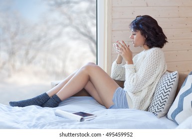 Young Beautiful Brunette Woman With Cup Of Coffee Sitting Home On The Bed By The Window. Winter Snow Landscape View. Lazy Day Off Concept