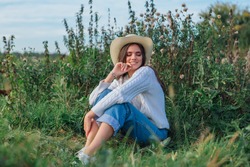 Young Beautiful Brunette Woman Dressed In A White Sweater, Jeans And Cowboy Straw Hat Sitting On Green Grass, Smiling And Laughing During Sunset