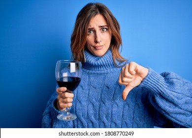 Young beautiful brunette woman drinking glass of red wine over isolated blue background with angry face, negative sign showing dislike with thumbs down, rejection concept