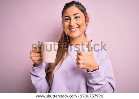 Young beautiful brunette woman drinking mug of coffee over isolated pink background happy with big smile doing ok sign, thumb up with fingers, excellent sign