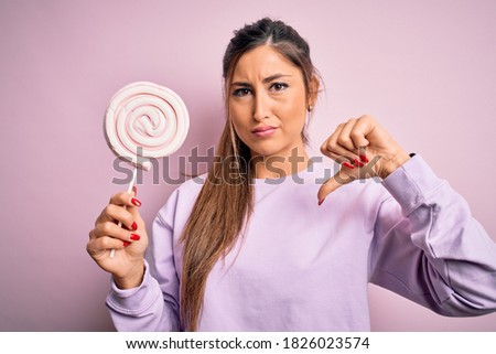 Young beautiful brunette woman eating sweet candy over isolated pink background with angry face, negative sign showing dislike with thumbs down, rejection concept