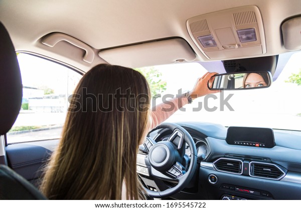 Young\
beautiful brunette woman is getting ready for a trip in a modern\
car. She is sitting on the front seat, looks and aligns the rear\
mirror on sunny light background. Travel\
concept
