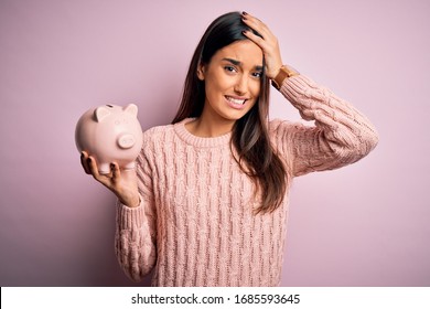 Young Beautiful Brunette Woman Holding Piggy Bank Saving Money For Retirement Stressed With Hand On Head, Shocked With Shame And Surprise Face, Angry And Frustrated. Fear And Upset For Mistake.