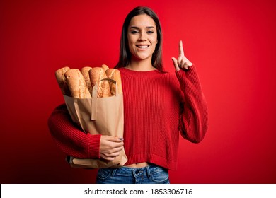 Young beautiful brunette woman holding paper bag with bread over red background surprised with an idea or question pointing finger with happy face, number one
